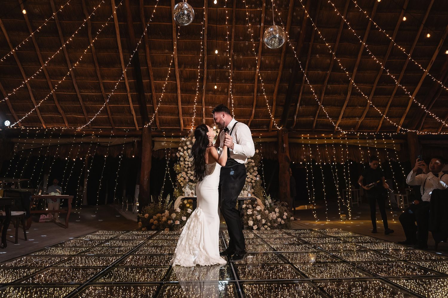 Newlyweds dancing beneath glowing string lights and mirror balls in a rustic barn reception.