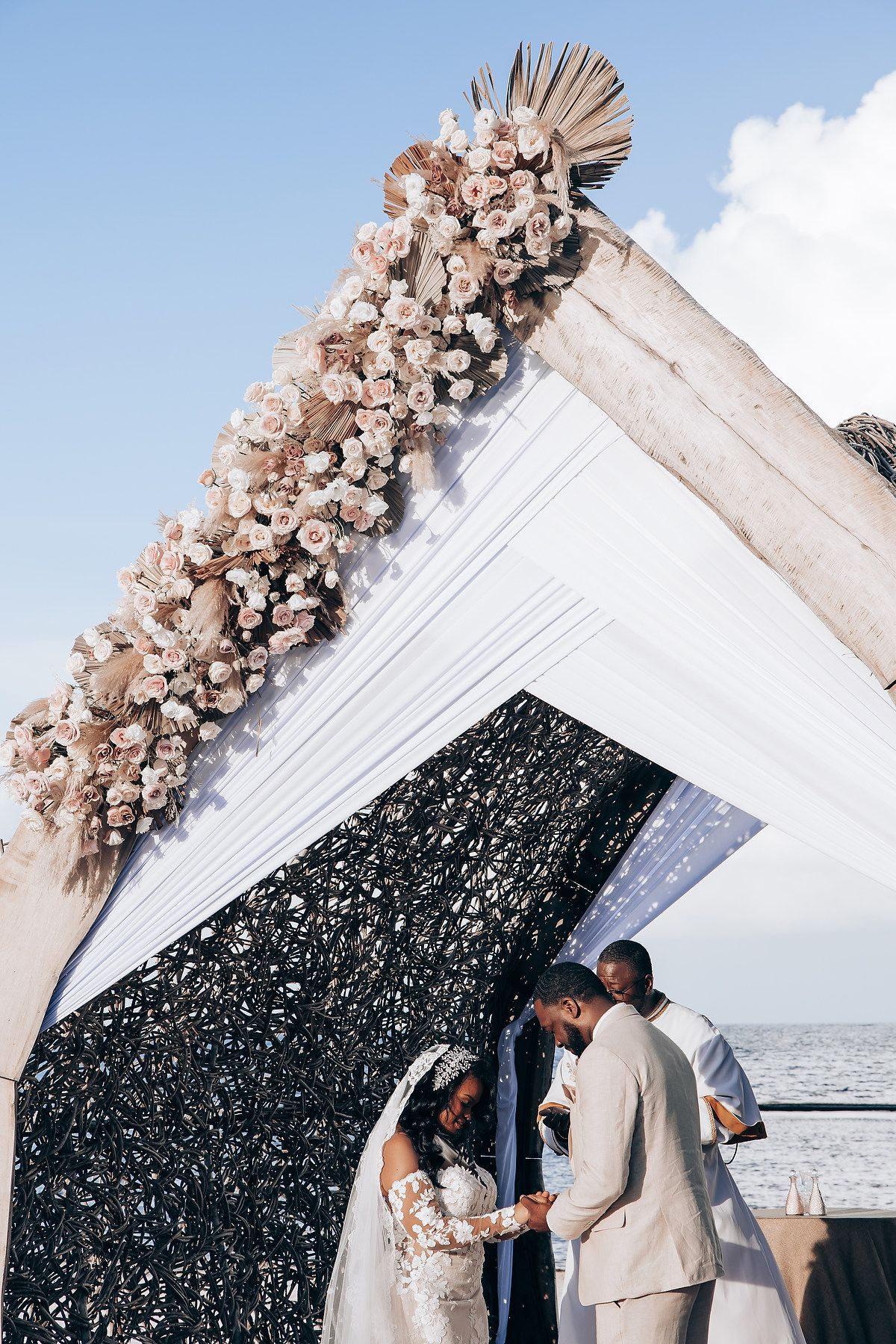 Intimate waterfront wedding ceremony under a decorated arch with pink flowers and black accent panel.