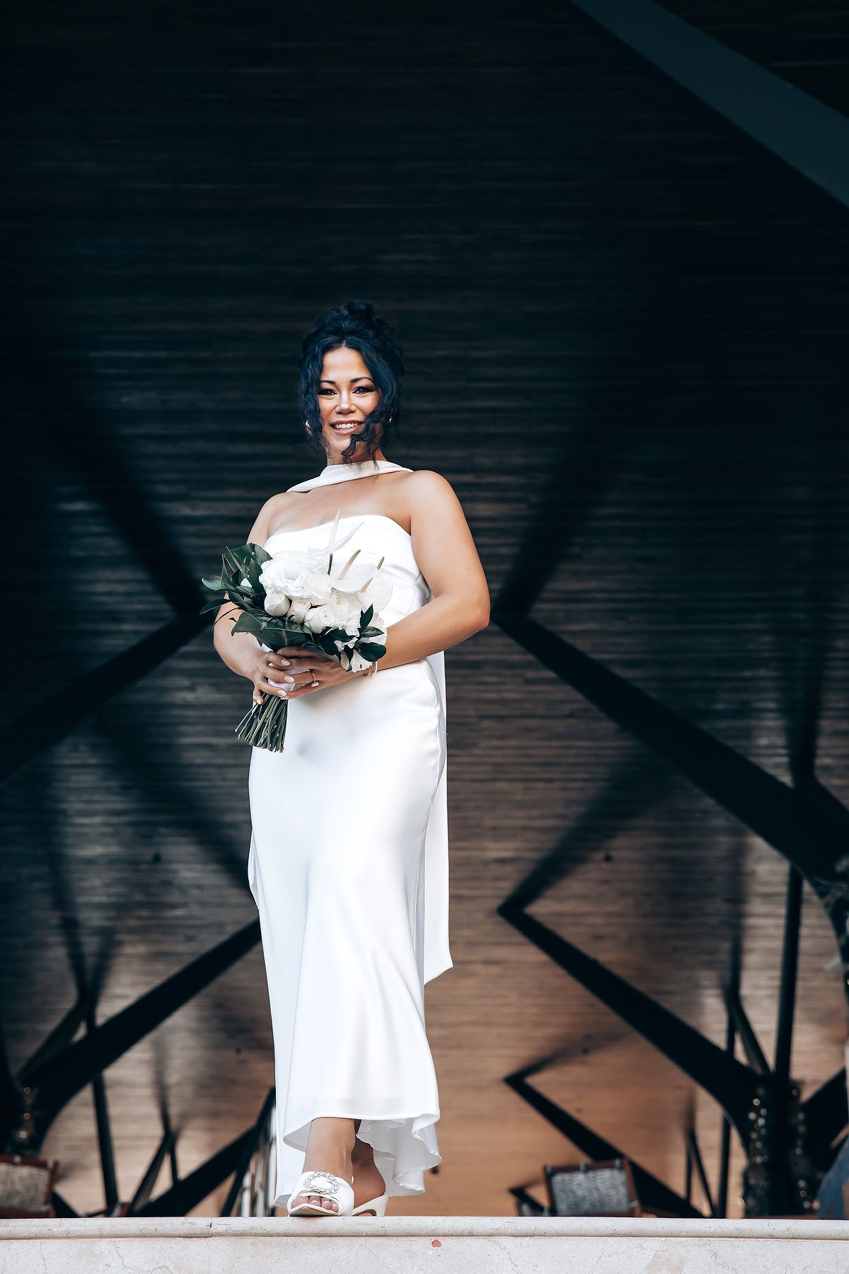 Person in white dress holding flowers stands against dark background with dramatic lighting.