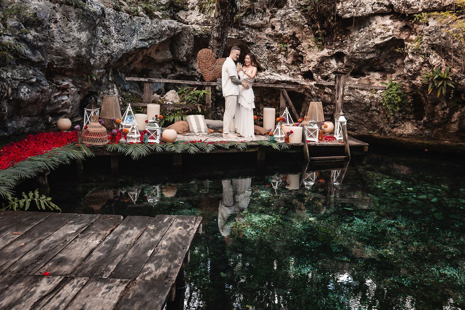 Romantic scene on wooden platform over emerald cenote waters with cave backdrop.