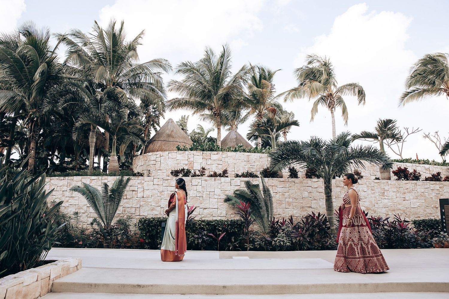 Two people in traditional Indian wedding attire standing on white steps surrounded by palm trees.