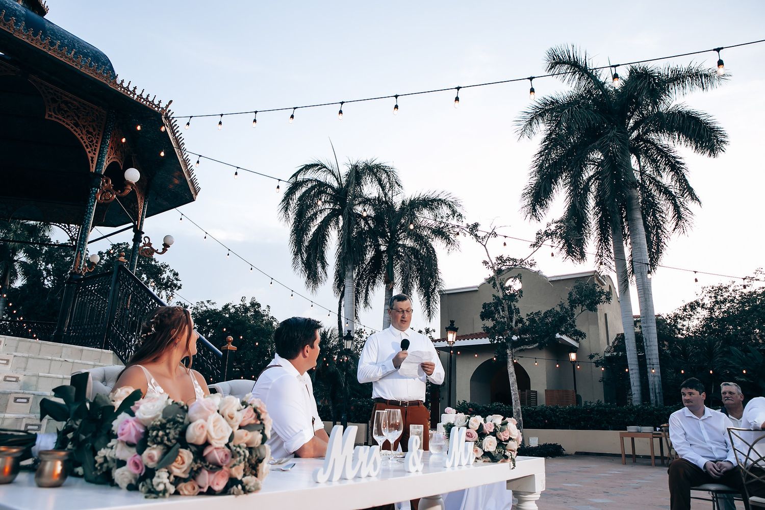 Outdoor wedding reception with string lights and palm trees at dusk.