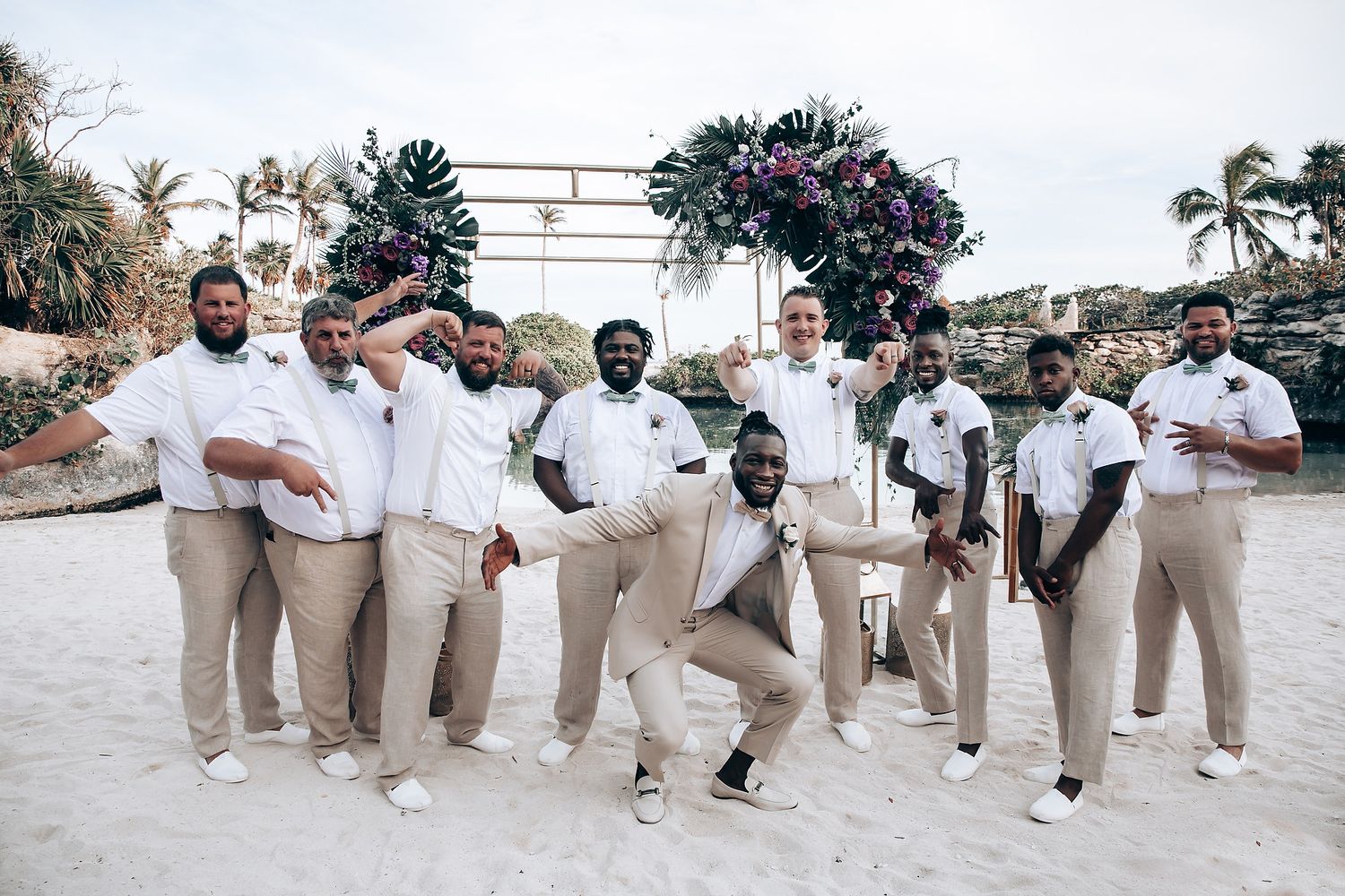 Groomsmen in beige suits and white shirts pose playfully for a group photo at a tropical venue.