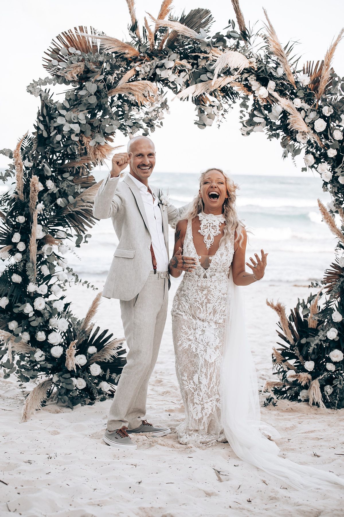 Happy couple sharing an exuberant moment under a tropical flower arch at their beach wedding.