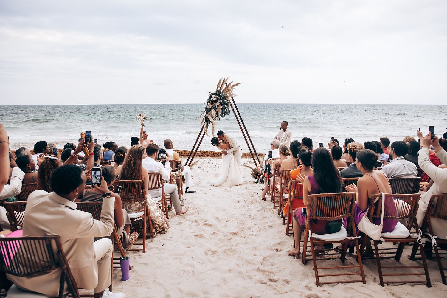 Beach wedding ceremony setup with guests watching couple under triangular arch by the ocean.