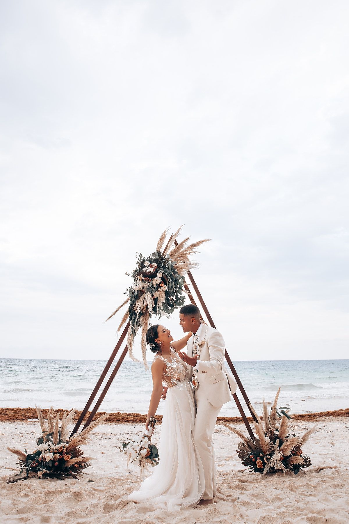 Intimate beach wedding ceremony with triangle arch and floral arrangements against ocean backdrop.