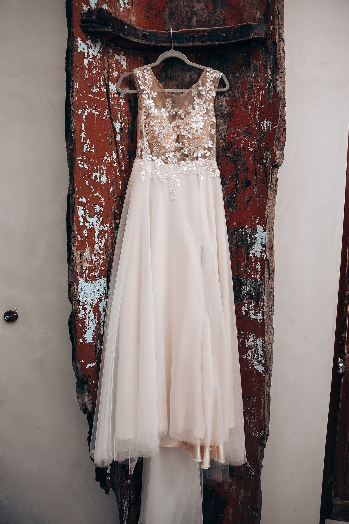 Delicate white lace wedding dress hanging against distressed red wooden wall.