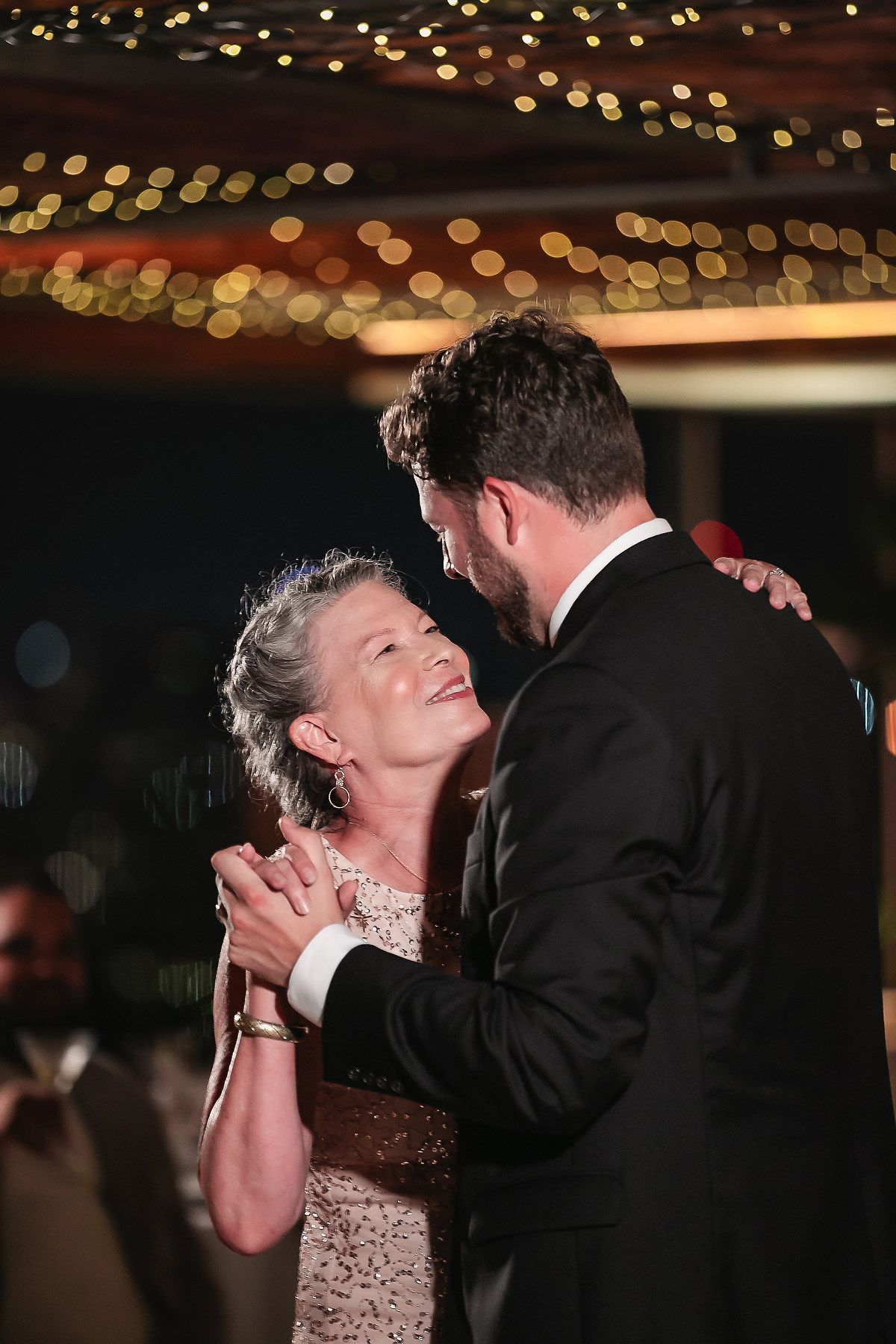 Couple dances beneath glowing string lights in a romantic reception venue setting.