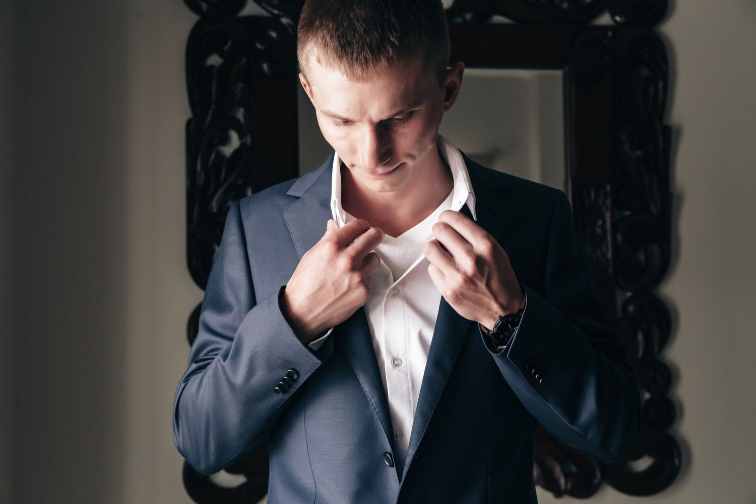 Groom in navy suit adjusts his collar in front of an ornate dark mirror before his wedding ceremony.