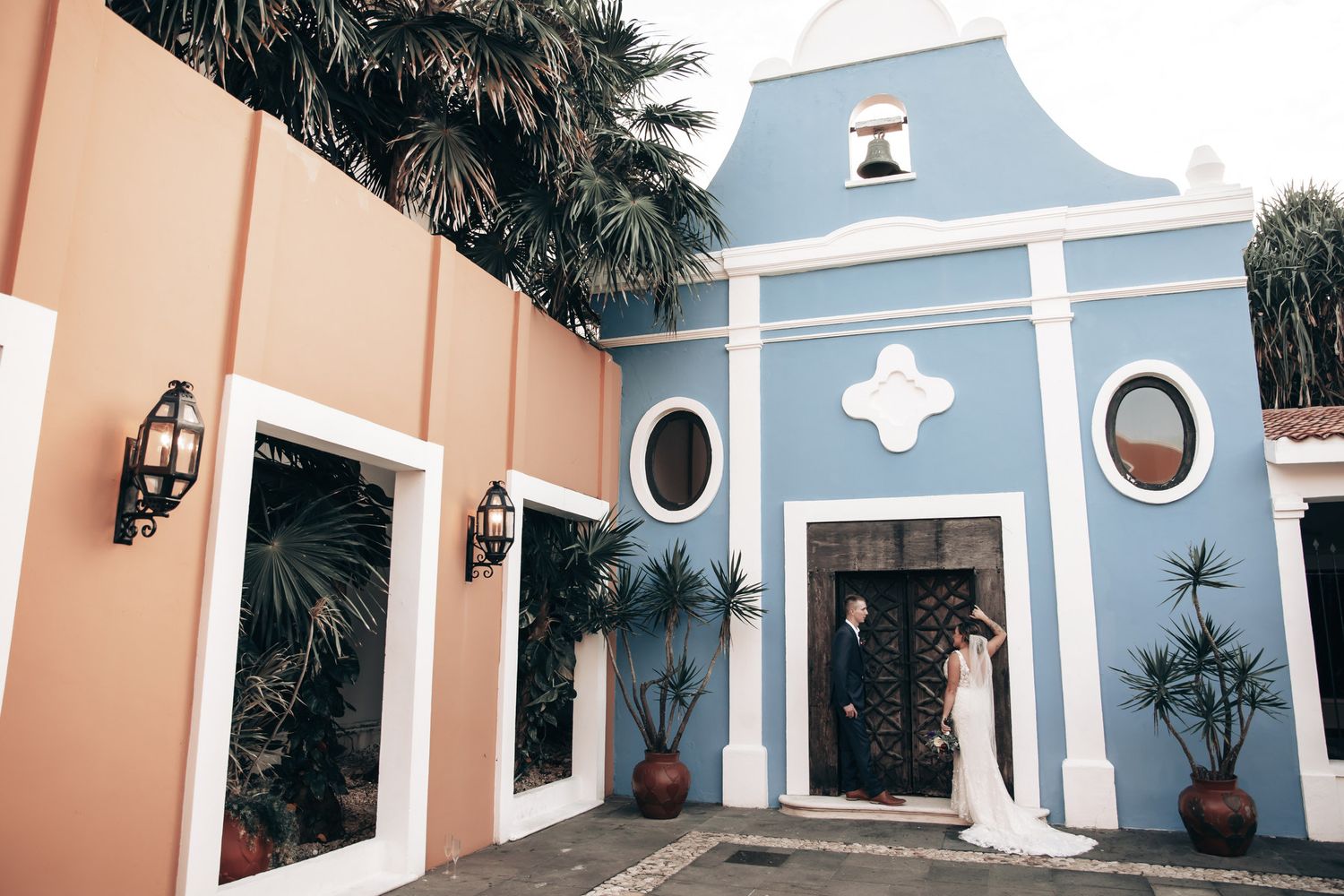 Bride and groom stand at entrance of colorful blue and pink chapel surrounded by tropical palms.