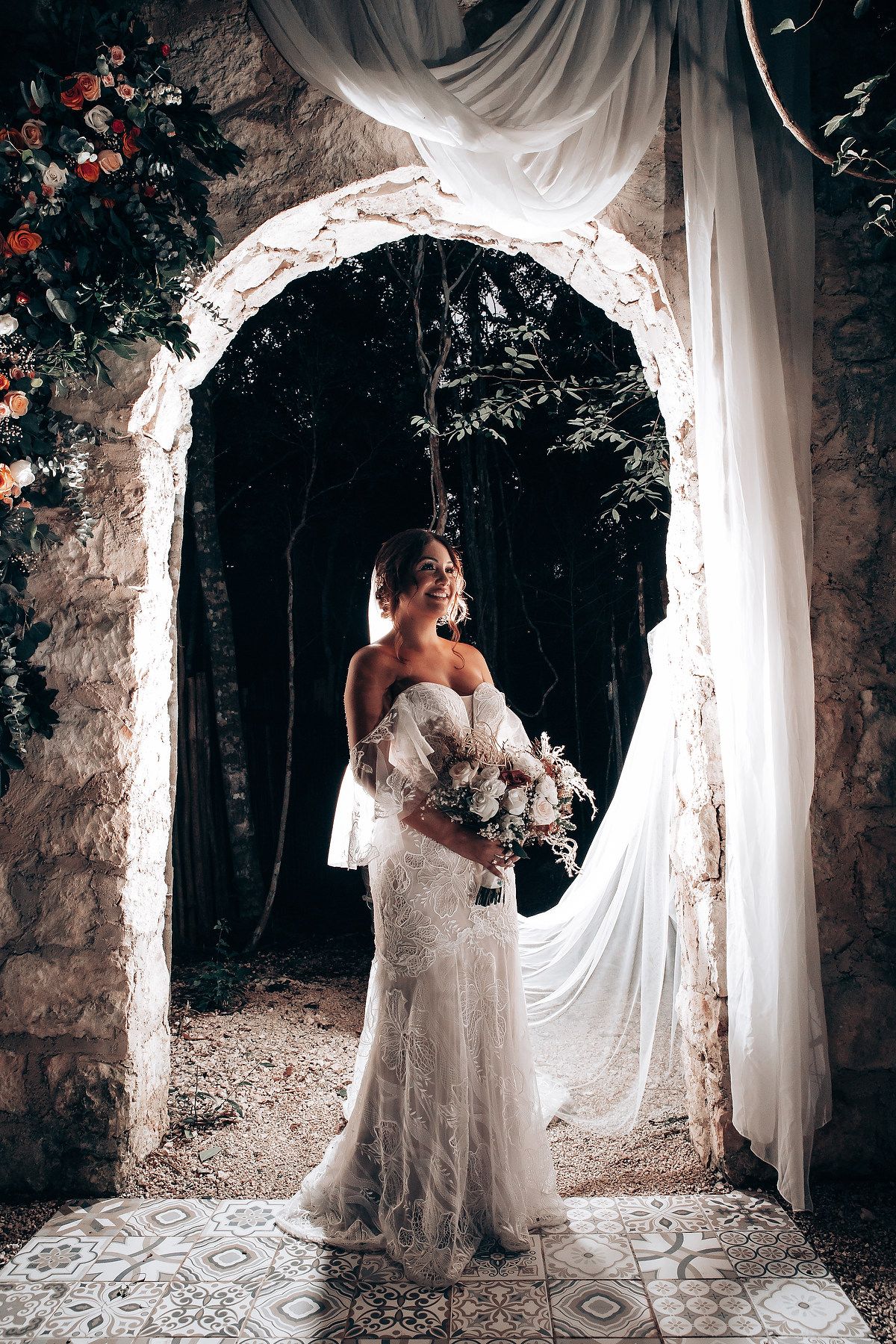 A bride in a lace gown poses under a romantic stone archway draped with white fabric and floral decorations.