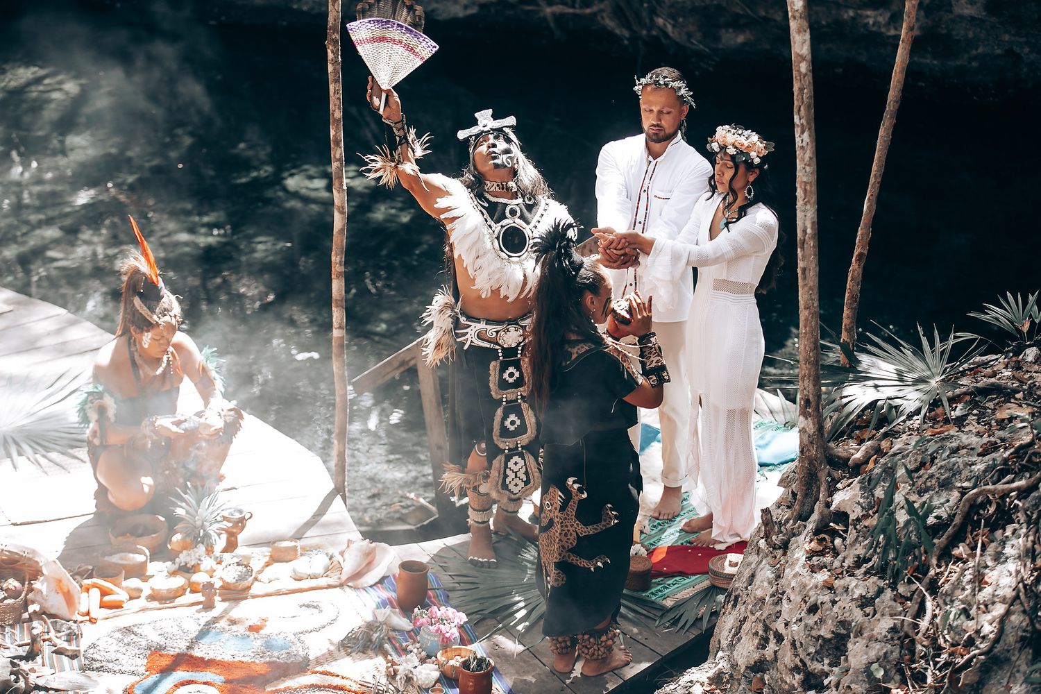 A traditional ceremony unfolds in a wooded area with participants dressed in white.