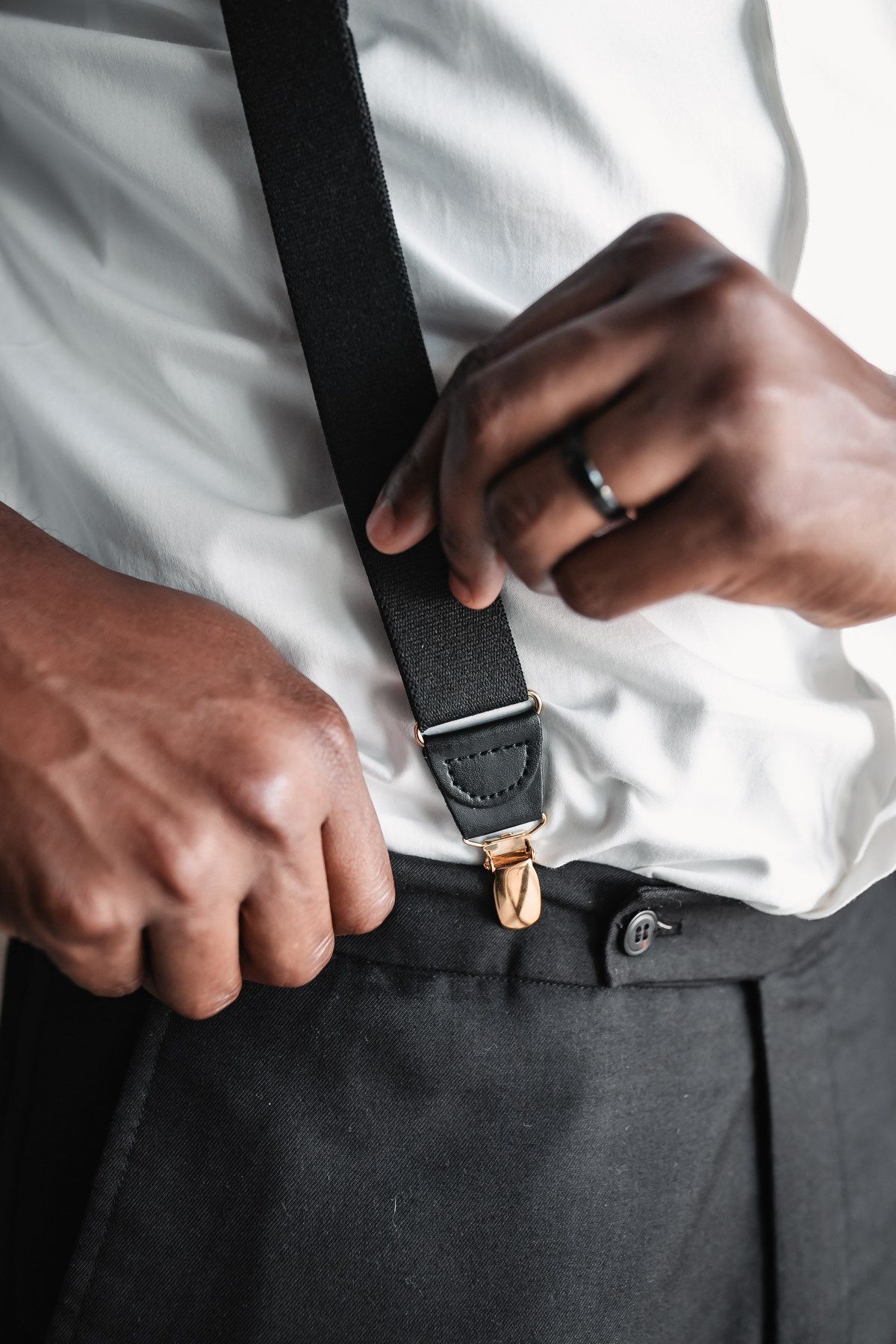 Groom adjusting black suspenders over white dress shirt with gold hardware detail.