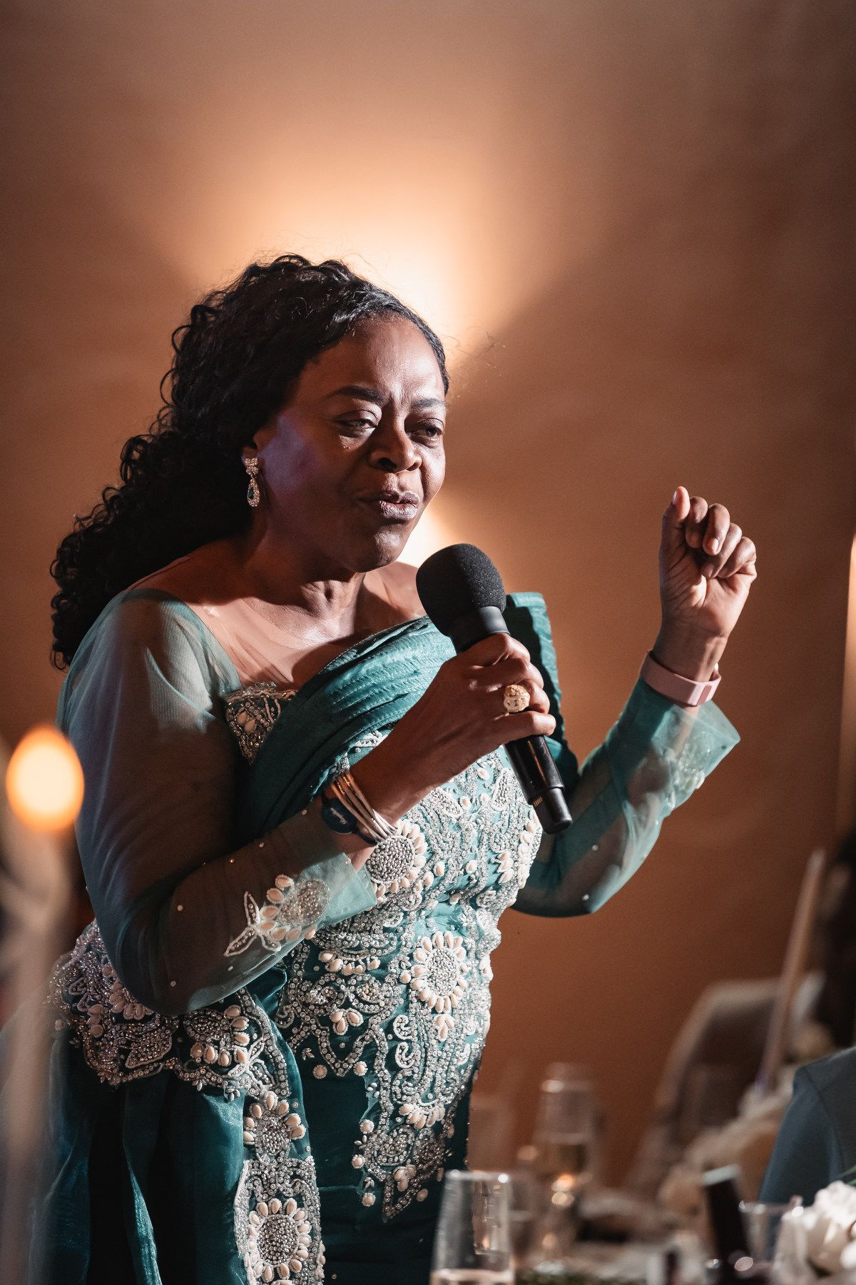 Elegantly dressed woman in teal lace gown delivers an emotional wedding speech holding a microphone.