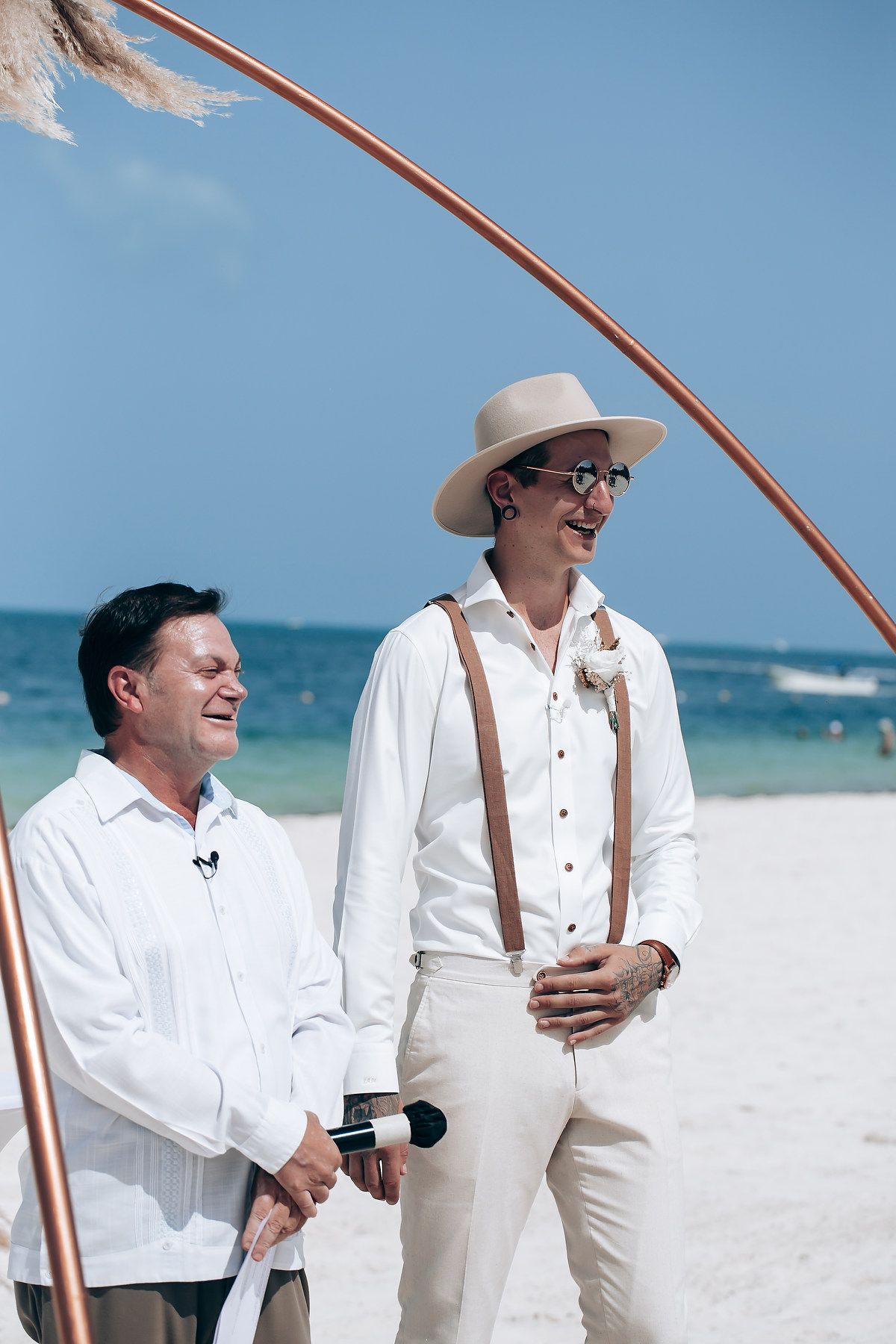 Two people in white outfits share a lighthearted moment on a sunny beach with ocean waves in the background.