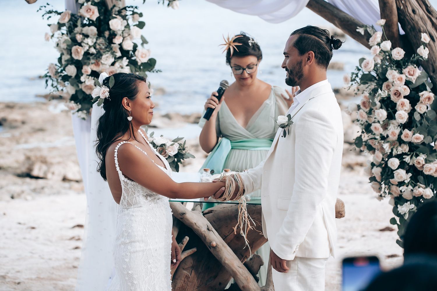Beachside wedding celebration with white floral arch and musicians performing.