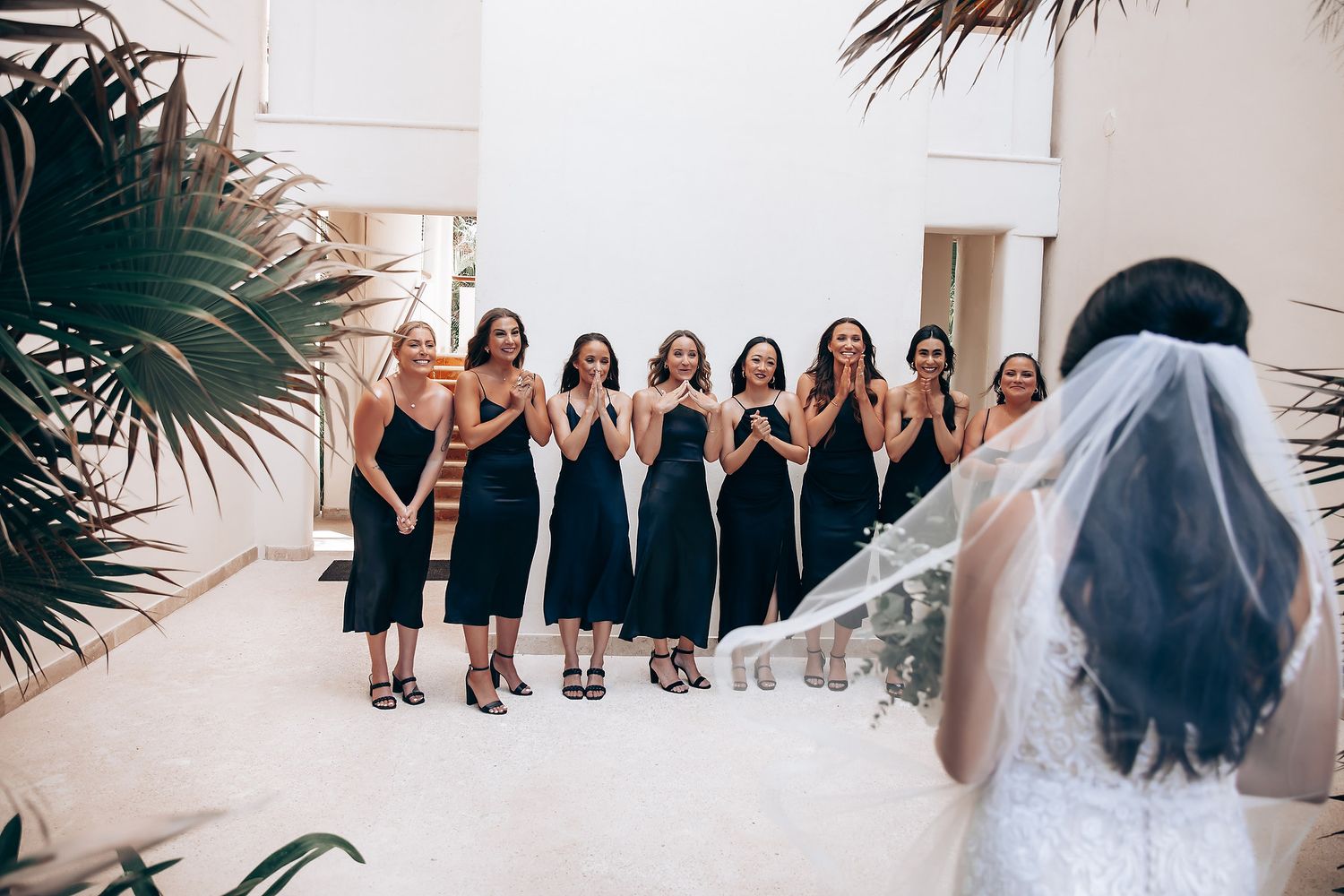 A group in black dresses reacts with excitement to a bride in her wedding gown.