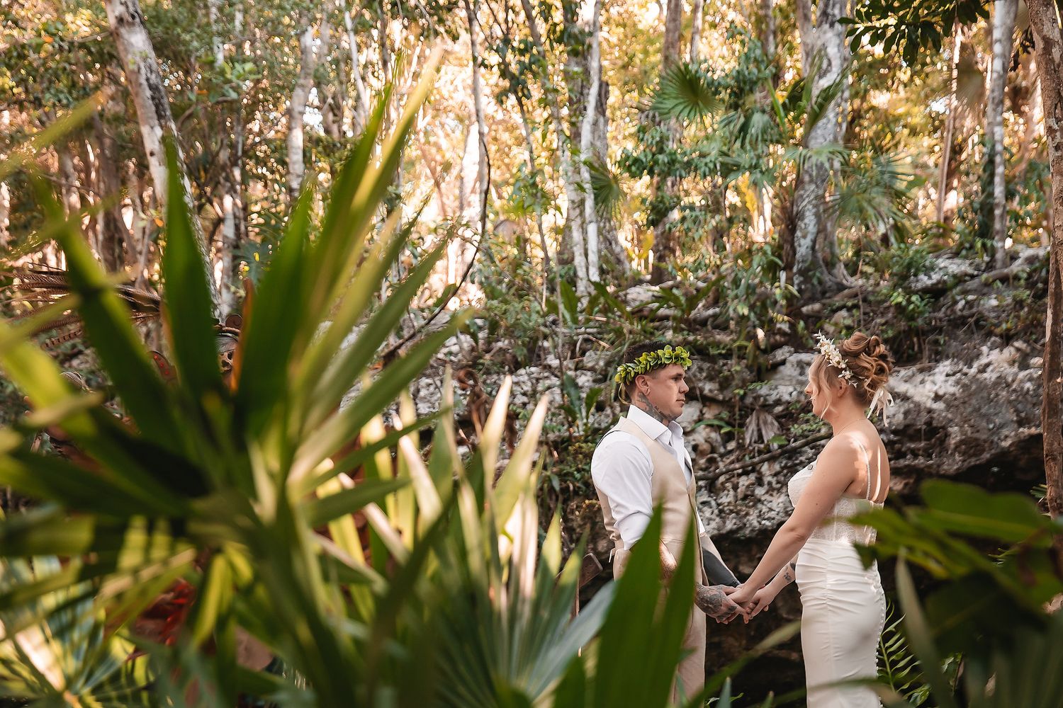Couple holds hands during outdoor wedding ceremony surrounded by lush tropical jungle greenery and ancient stone ruins.