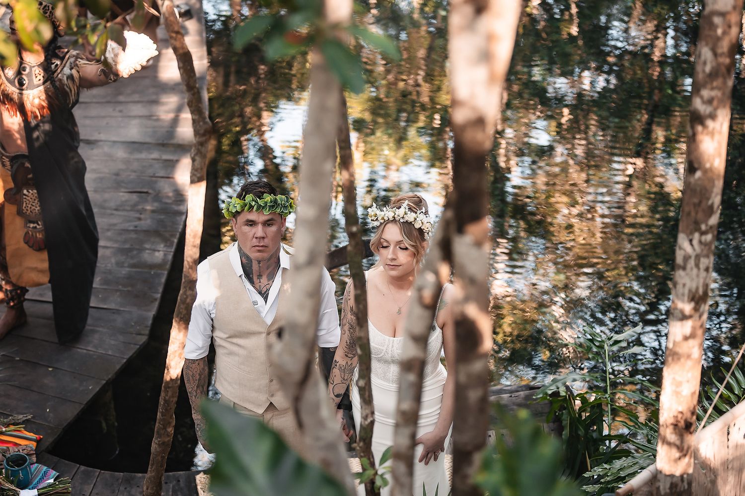 Bride and groom in floral crowns walk through lush woodland setting near water at outdoor wedding ceremony.