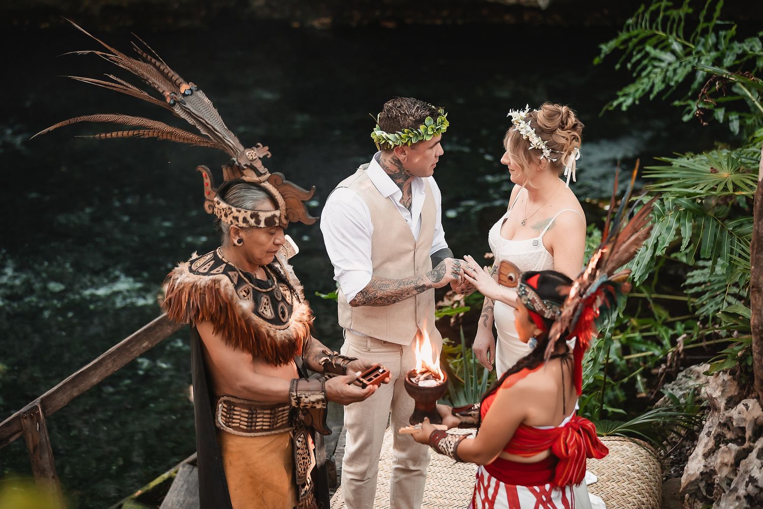 Couple exchanges vows in Mayan ceremony with costumed officiant holding fire near cenote in jungle.