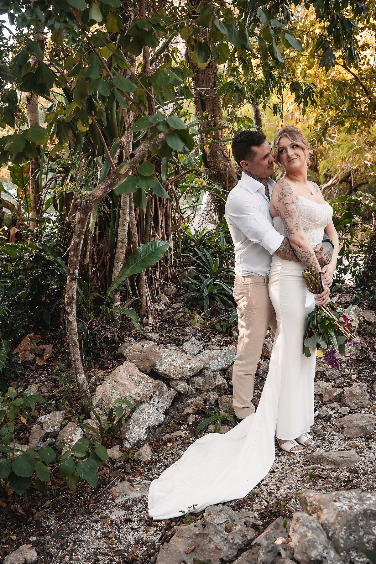 Bride and groom embrace among lush tropical greenery and rocks during outdoor wedding photo session.