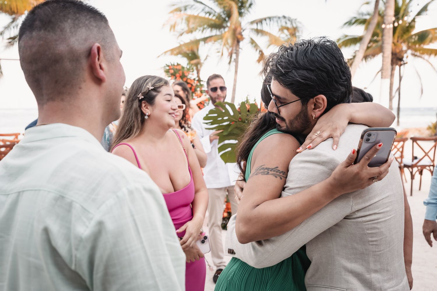 Wedding guests hugging and celebrating at tropical beach reception with palm trees at sunset.