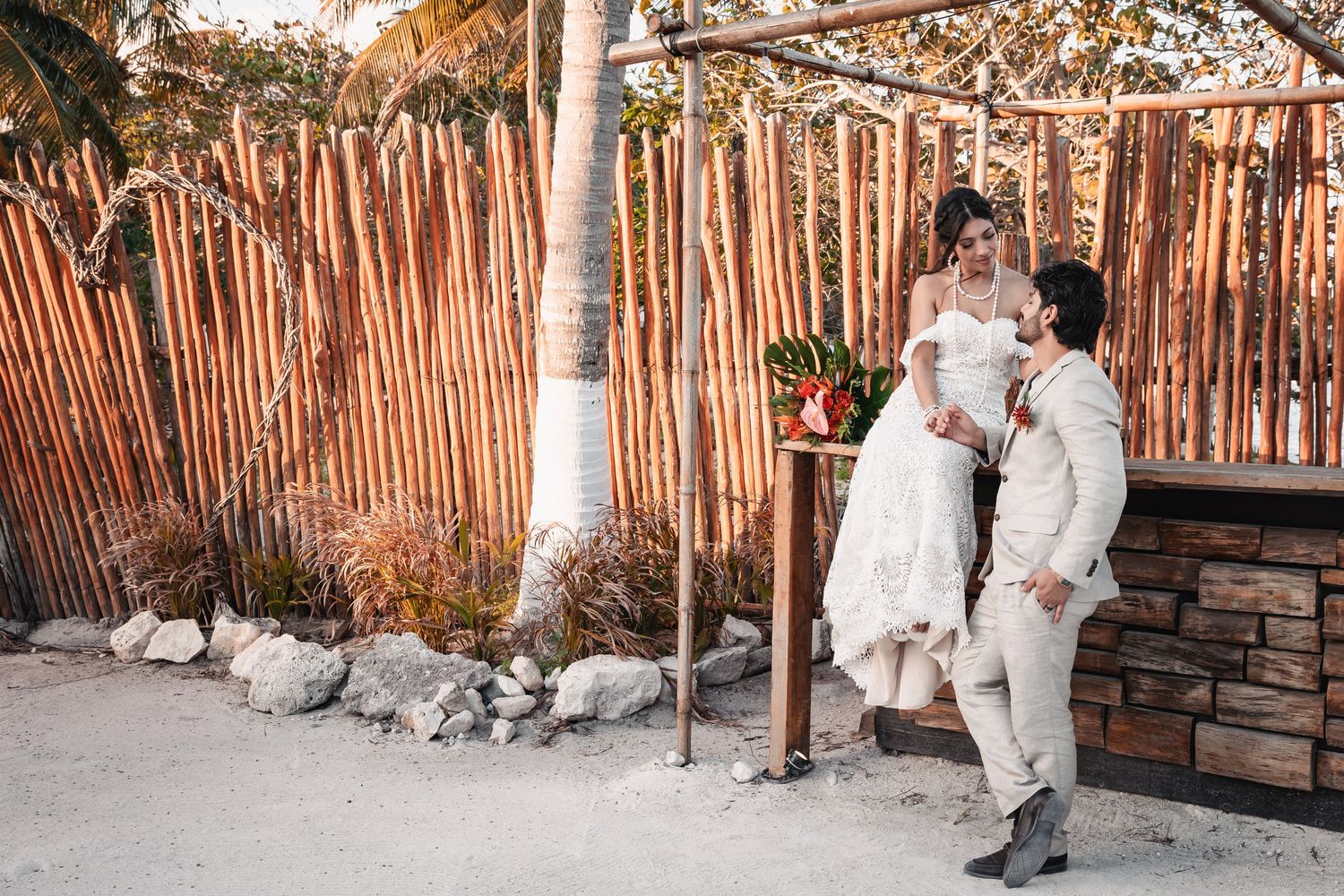 Bride and groom posing together near bamboo fence at sunset beach venue.