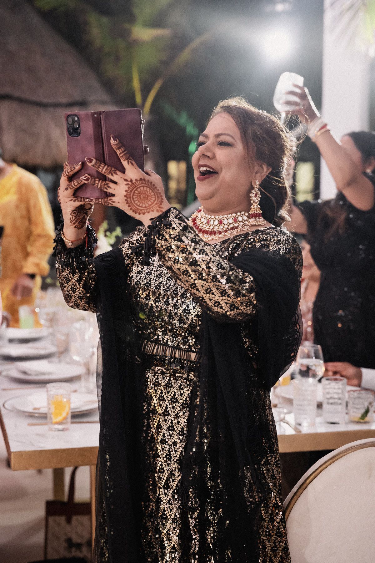 Woman in black gold embroidered lehenga with mehndi hands laughing while taking selfie at wedding reception.