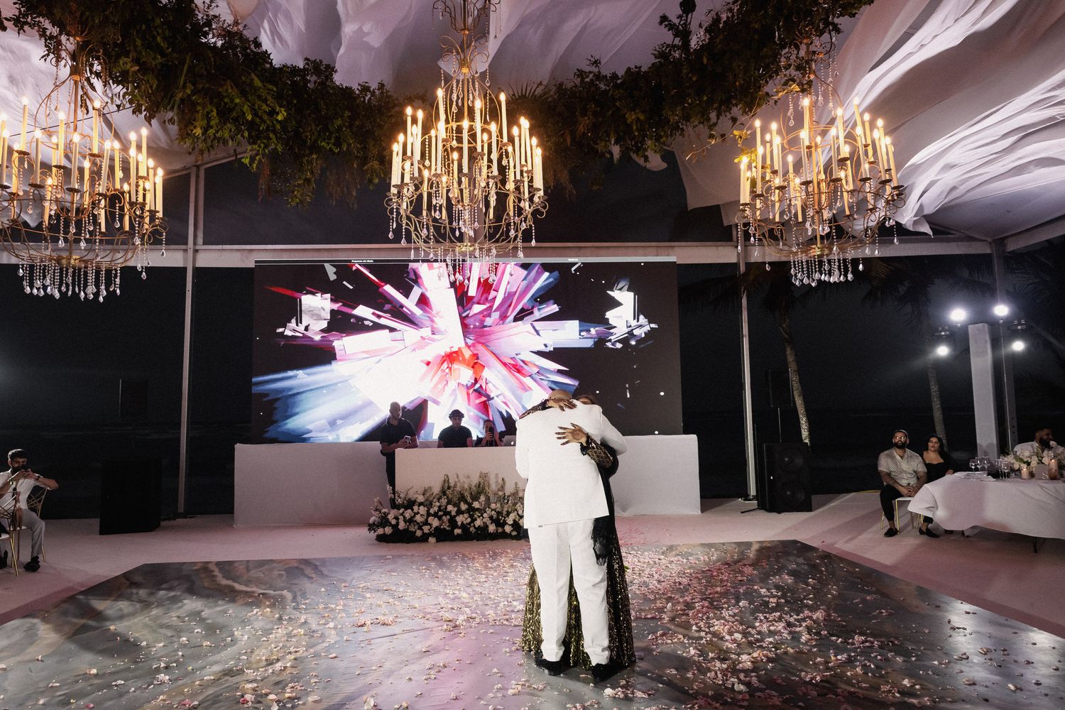 Newlyweds embracing on decorated dance floor under chandeliers with fireworks display backdrop.