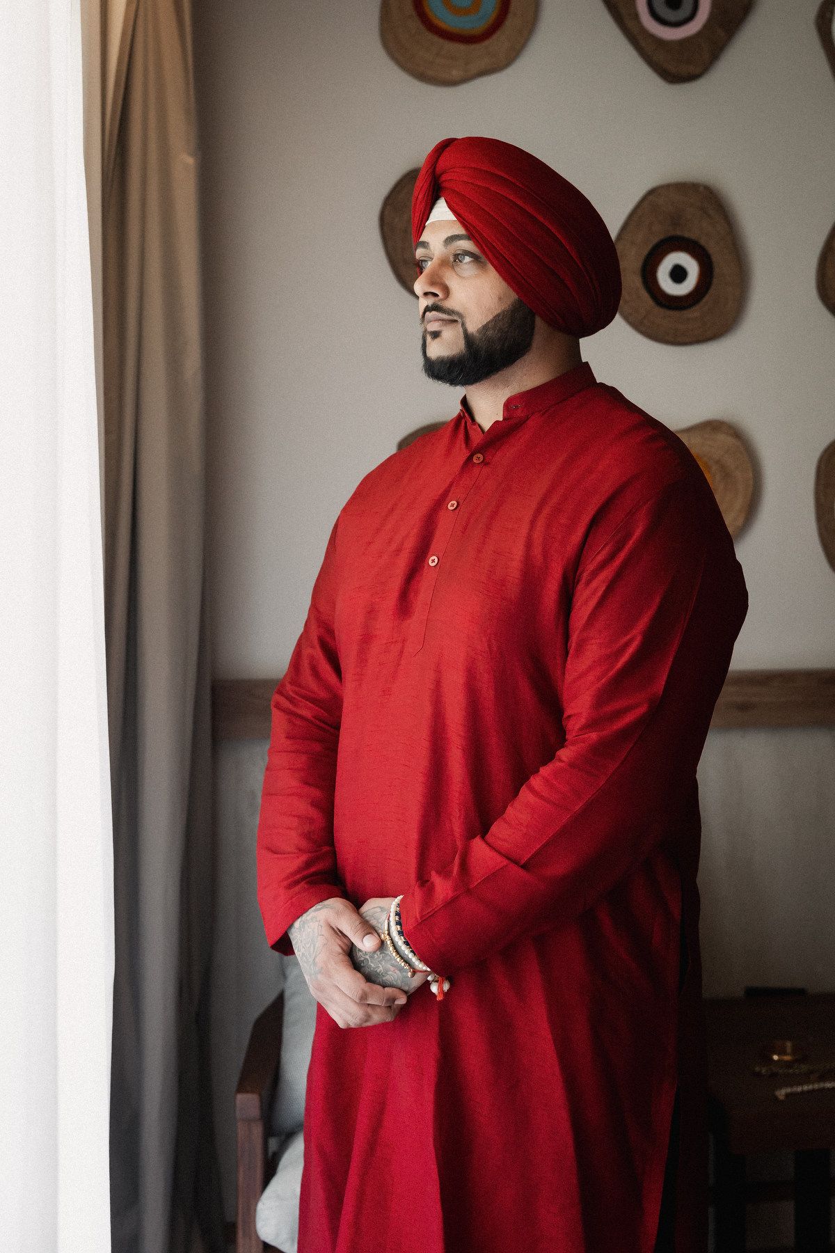 A Sikh groom in a red kurta and turban stands elegantly indoors before his wedding ceremony.