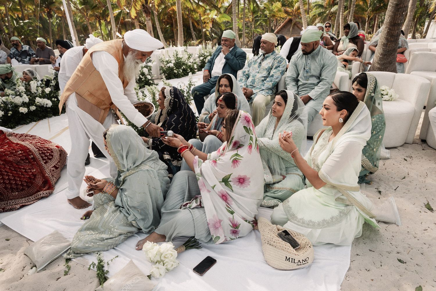 Guests in traditional Indian attire gather on sandy beach during Sikh wedding ceremony outdoors.