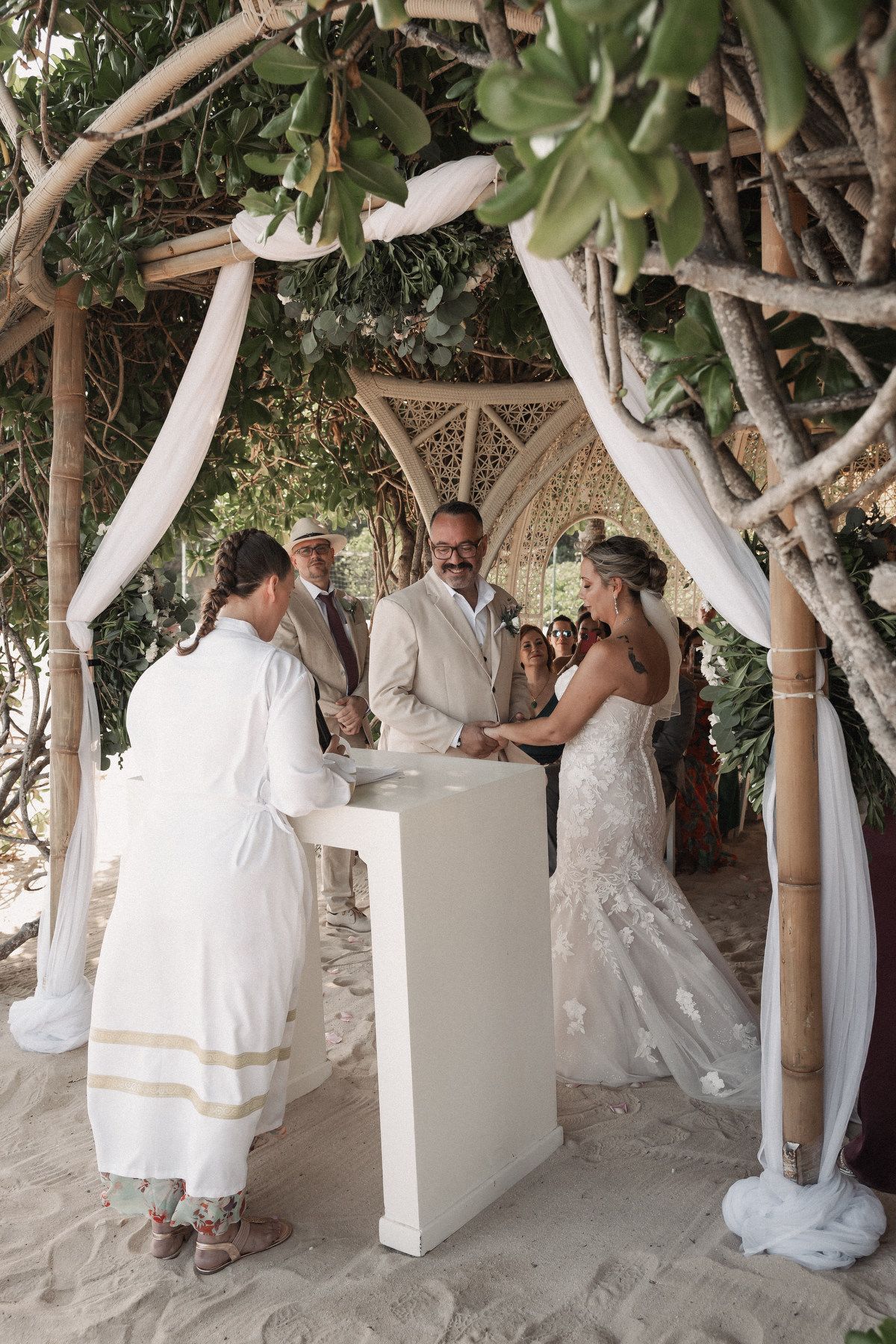 Bride and groom stand at altar under rustic wooden arch on beach during outdoor wedding ceremony with officiant.