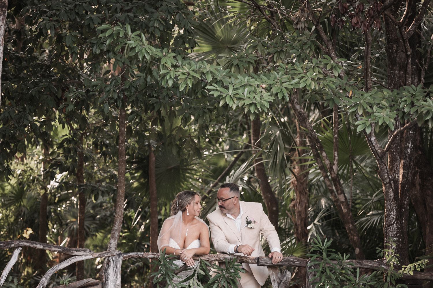 Bride and groom sit together laughing among lush green trees in natural setting.
