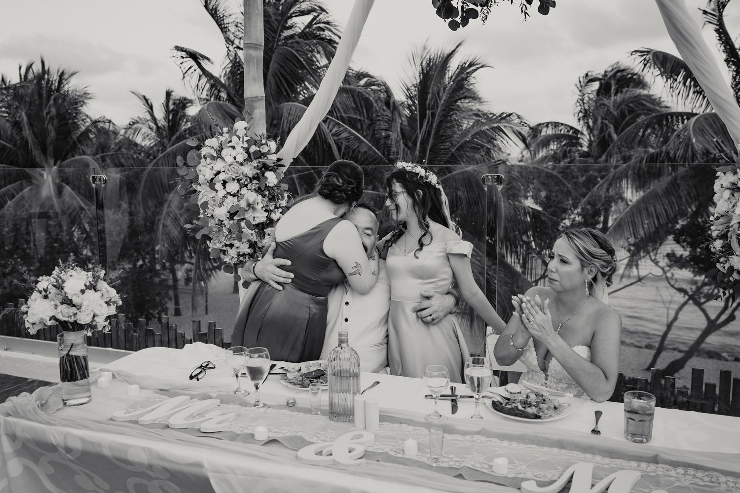 Black and white photo of bride sharing emotional embrace with guests at tropical outdoor reception.