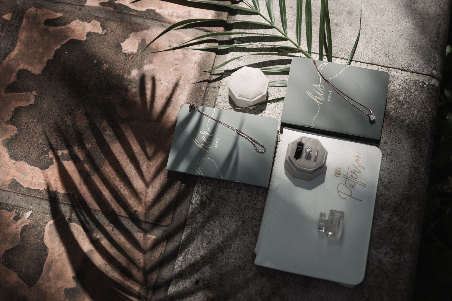 Flat lay of elegant wedding jewelry and accessories on a gray surface with tropical palm leaf shadow.