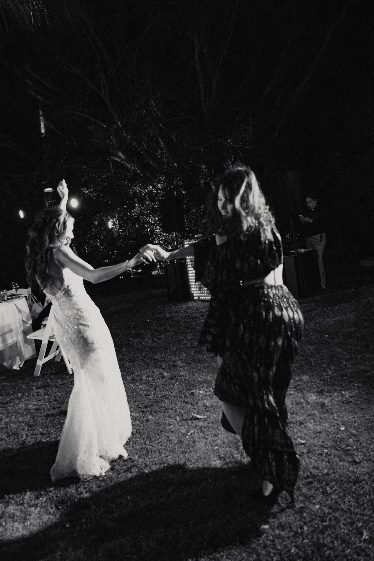 Black and white photo of bride dancing joyfully with female guest at outdoor nighttime wedding reception.