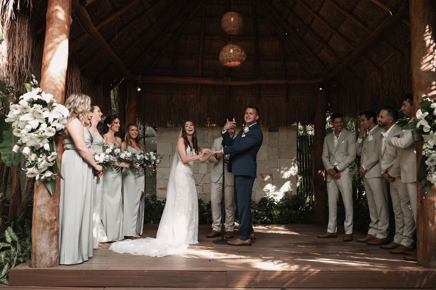 Bride and groom exchange vows under thatched roof with bridesmaids in sage green and groomsmen in tan.