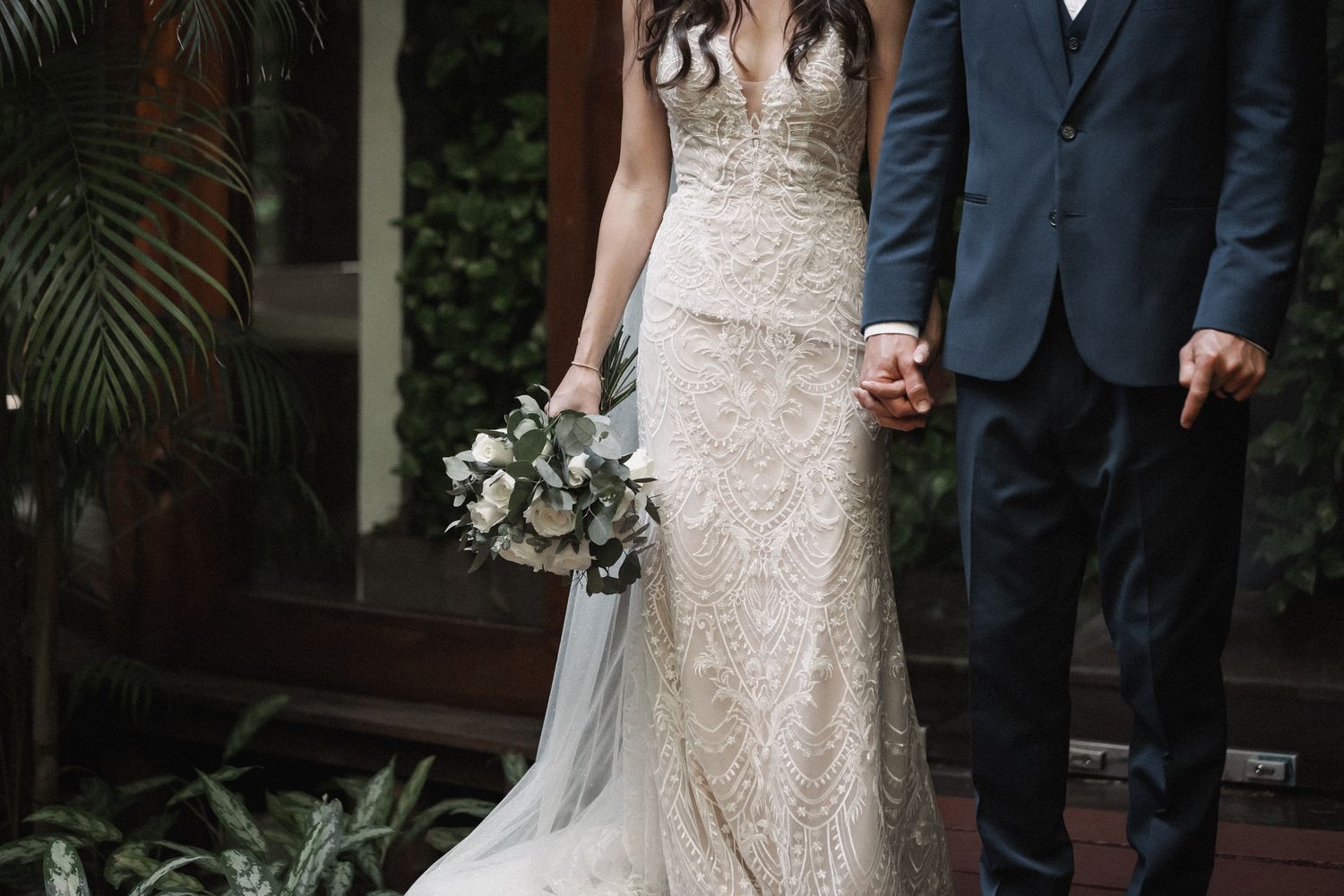 Bride in lace gown holds bouquet while holding hands with groom in navy suit.