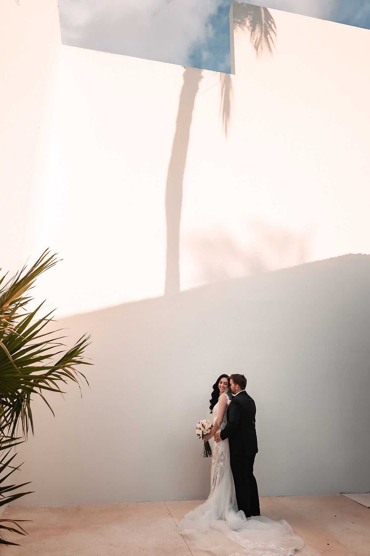 Bride and groom pose together against a white wall with palm tree shadows at a sunny outdoor wedding venue.