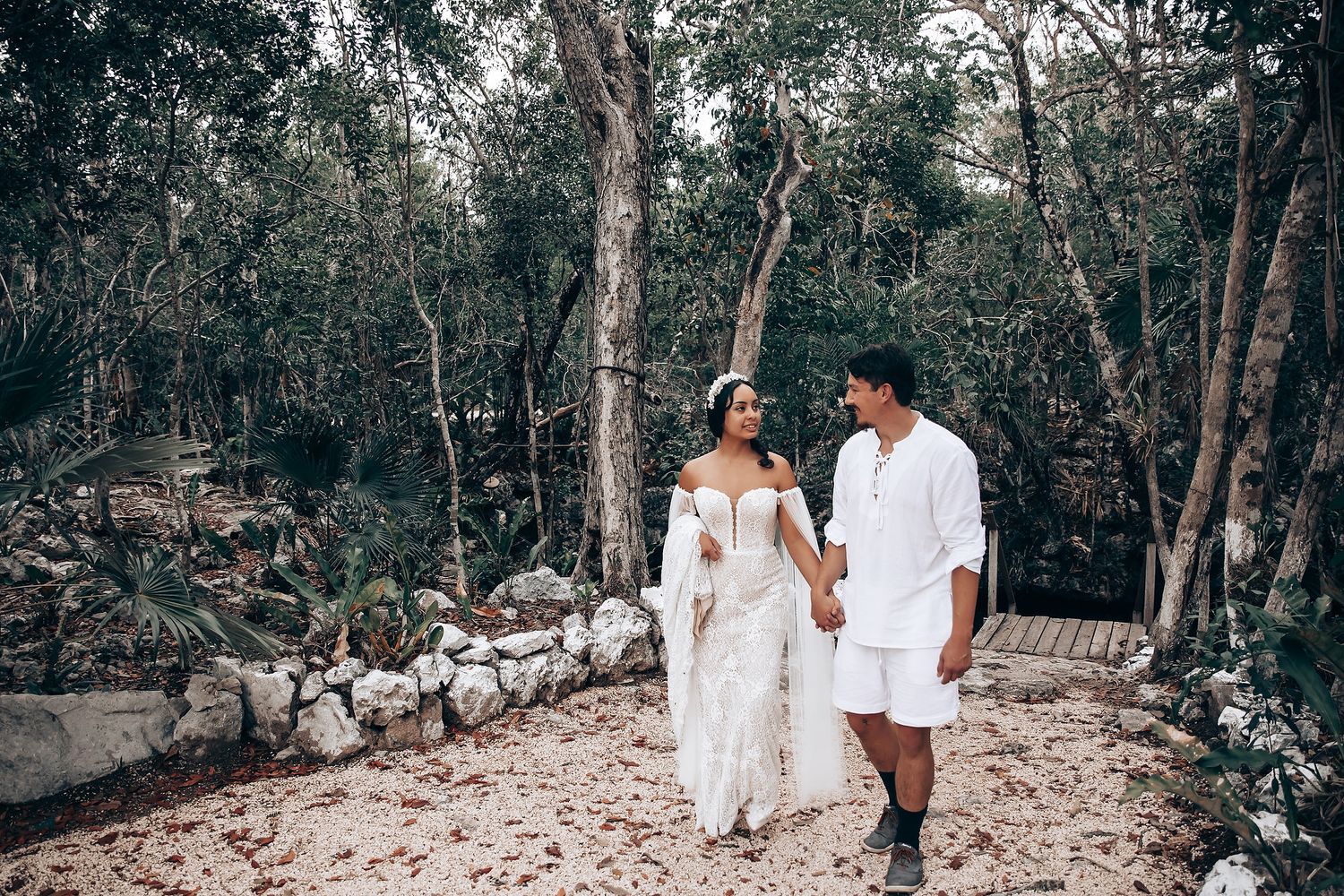 A couple in white attire walks together through a wooded area during a romantic photoshoot.