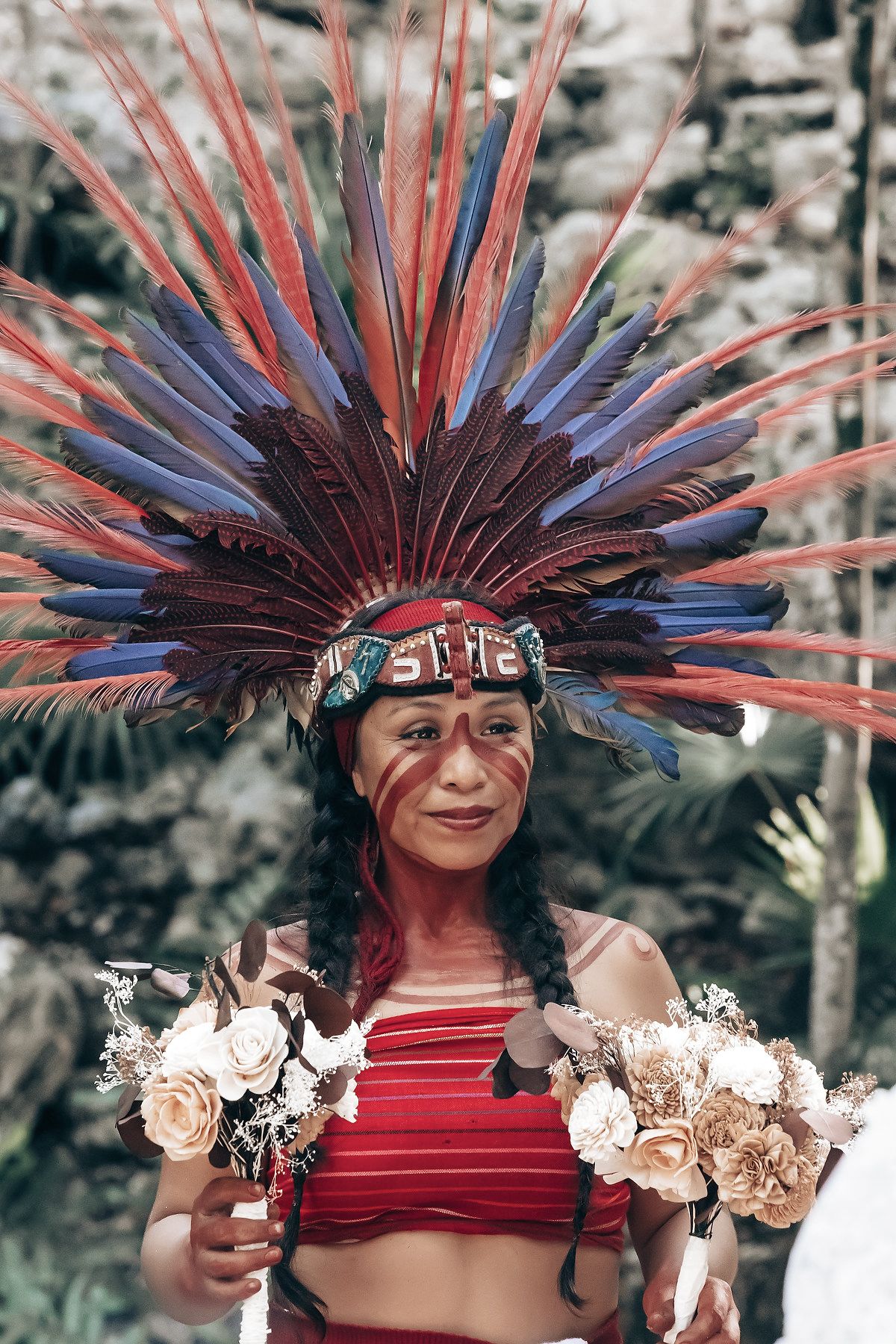 A woman in a red and black headdress and costume standing in a forest with trees and foliage.