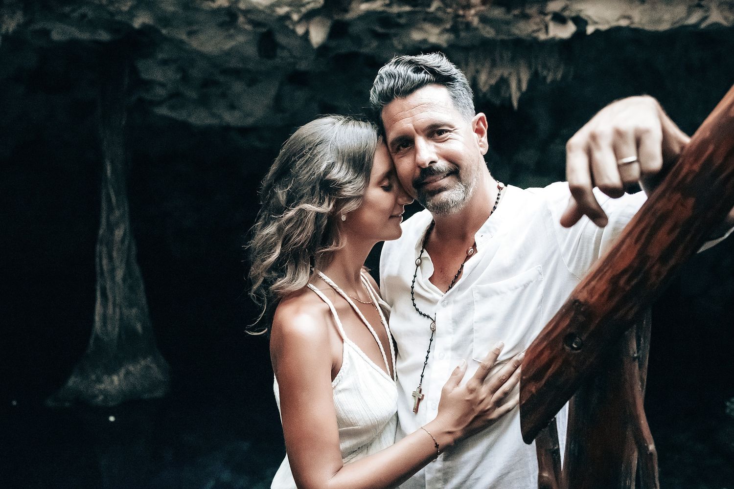 A couple in white clothing embraces in a natural, forested setting.