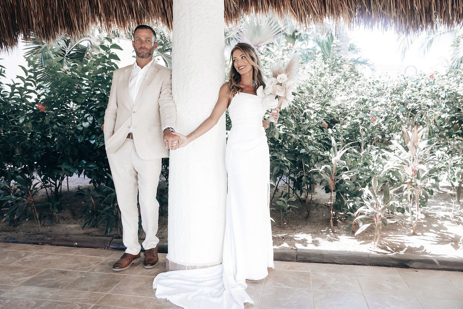 A couple in formal attire stand together in a tropical setting.