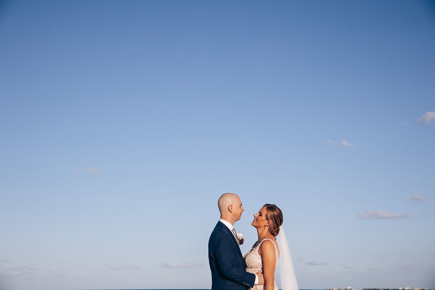 A bride and groom stand together against a blue sky backdrop.