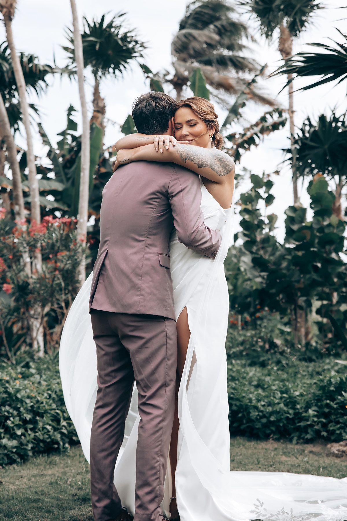 Couple embracing in tropical garden setting with palm trees and white wedding attire.