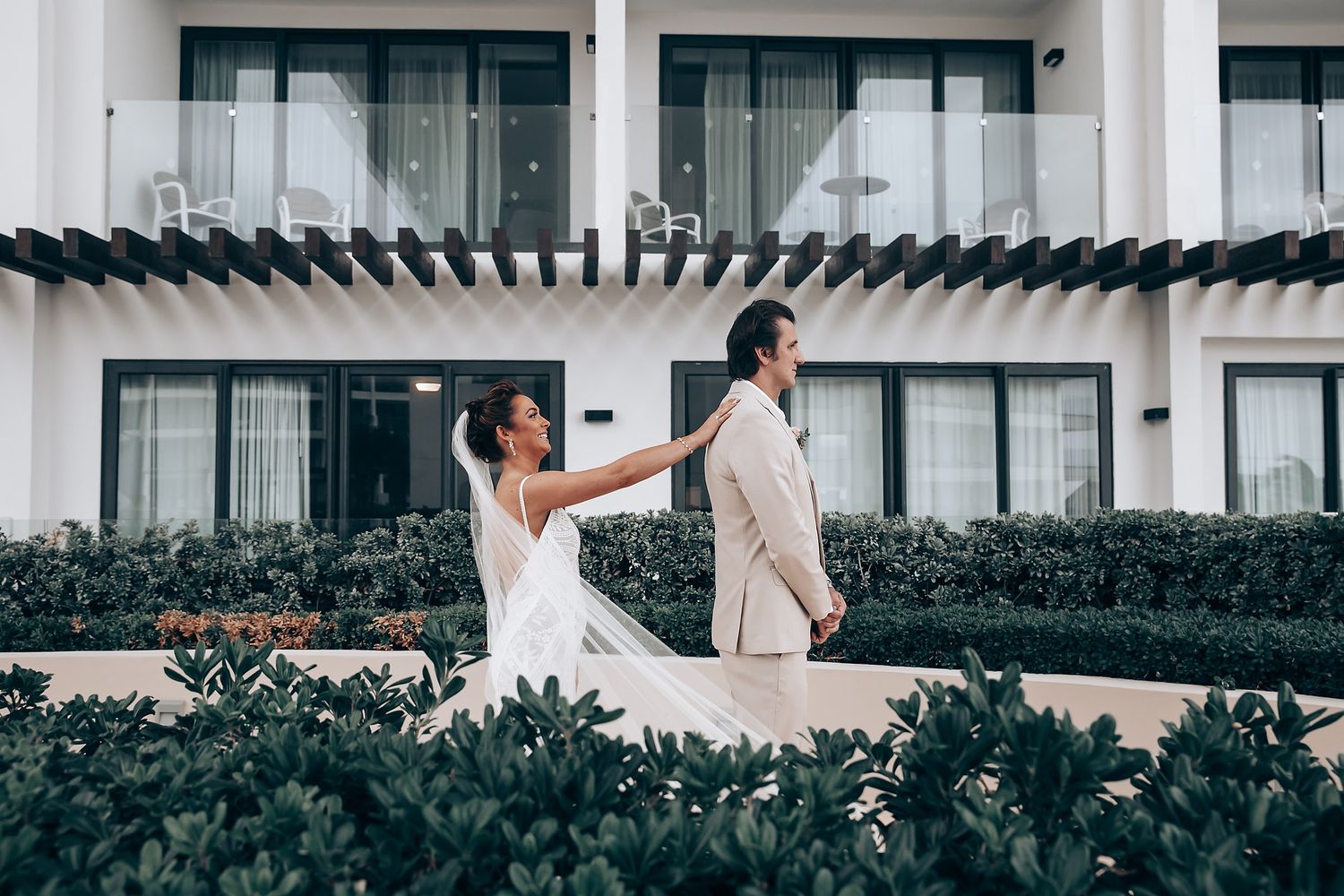 Two people in wedding attire walk along a path bordered by green hedges outside a modern white building.