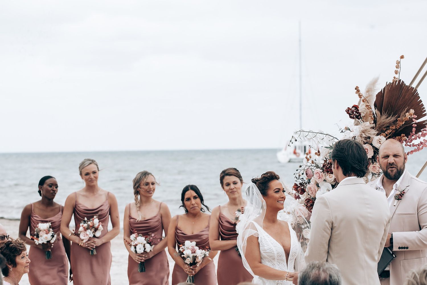 Wedding party lines up on beach under white geometric arch with floral decorations.