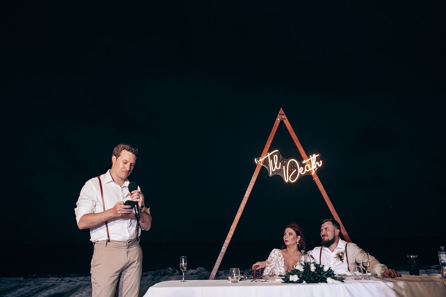 Evening beach wedding reception with string lights and wooden arch structures.