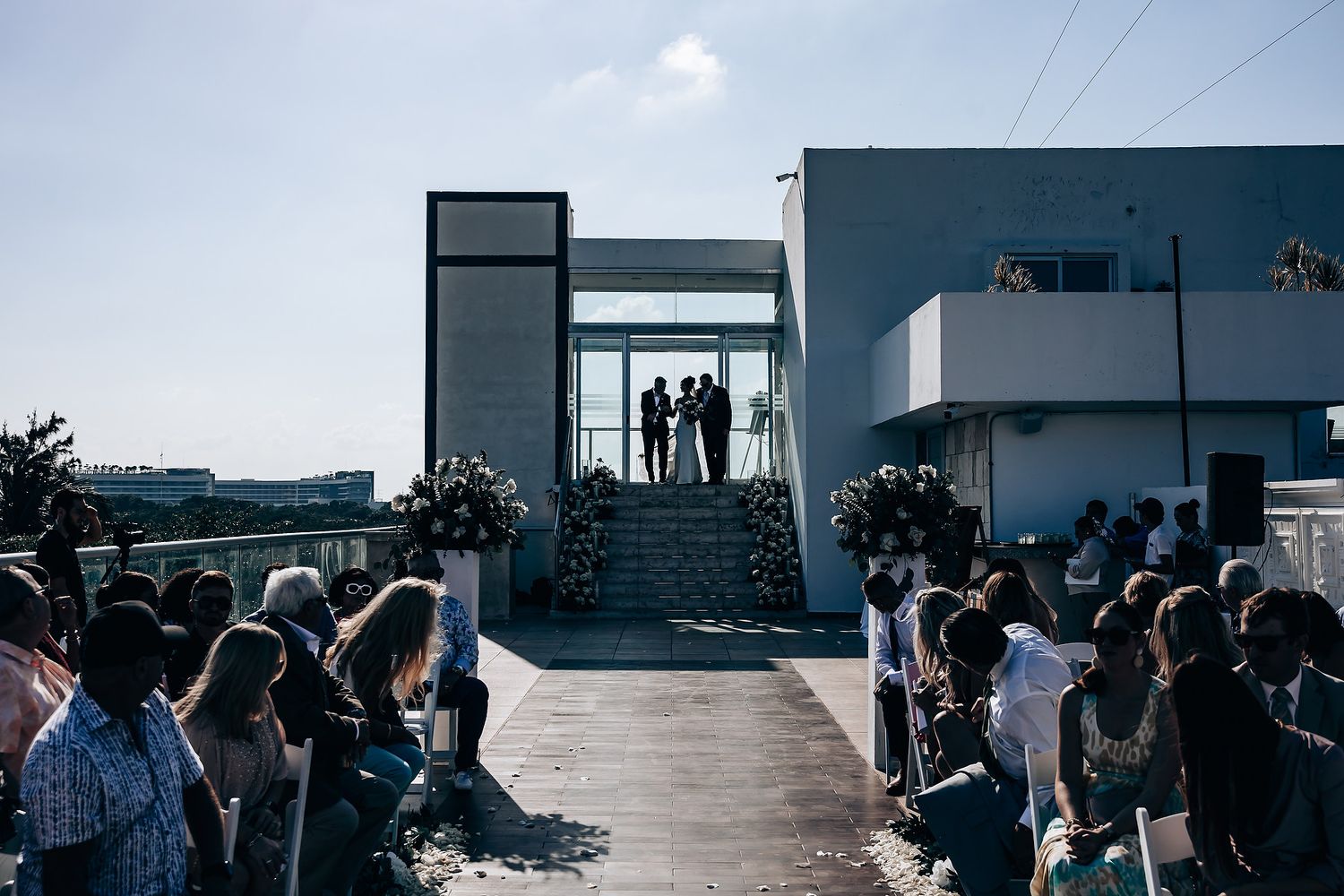 Wedding guests seated in white chairs at an outdoor ceremony overlooking the ocean on a sunny day.