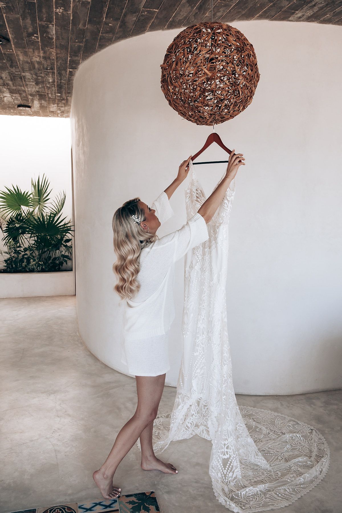 Woman in white dress reaching up to adjust a hanging brown woven light fixture in a minimalist interior space.